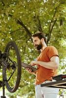 jong Kaukasisch mannetje innemend in fiets onderhoud vakkundig vaststelling gebroken wiel met de steun van zijn telefoon tablet. energiek en enthousiast Mens ijverig repareren fiets componenten gebruik makend van digitaal foto