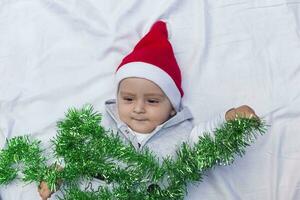 weinig grappig de kerstman jongen spelen met Kerstmis of nieuw jaar vakantie feestelijk guirlande. 1 jaar oud baby jongen in de kerstman claus kap. foto