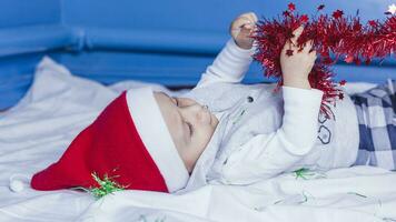 weinig grappig de kerstman jongen spelen met Kerstmis of nieuw jaar vakantie feestelijk guirlande. 1 jaar oud baby jongen in de kerstman claus kap. foto