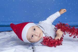 weinig grappig de kerstman jongen spelen met Kerstmis of nieuw jaar vakantie feestelijk guirlande. 1 jaar oud baby jongen in de kerstman claus kap. foto