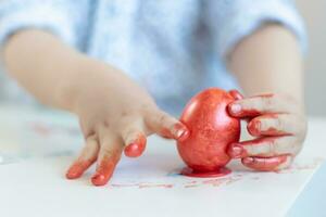 een kind zet een rood Pasen ei Aan een staan met zijn handen gebrandschilderd met verf Aan een wit tafel. foto