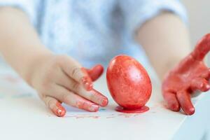 een kind zet een rood Pasen ei Aan een staan met zijn handen gebrandschilderd met verf Aan een wit tafel. foto
