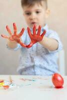 de jongen looks Bij zijn handen besmeurd en gebrandschilderd met rood verf en Aan de tafel Aan een staan is een rood Pasen ei. foto