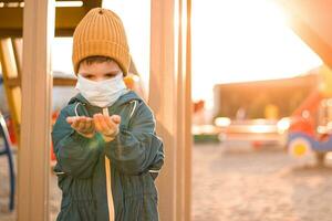 een jongen in een beschermend masker Toneelstukken in de speelplaats en morsen zand van zijn handen gedurende de pandemisch van coronavirus en covid - 19 foto