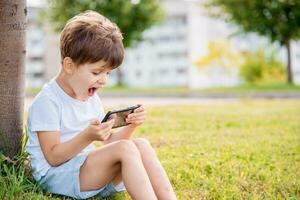 vrolijk kind zittend Aan de gras looks tekenfilms in de telefoon in de zomer Bij zonsondergang. schattig jongen hebben pret in natuur foto