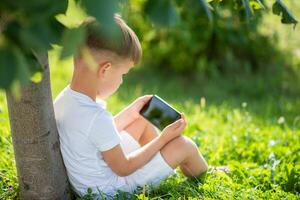 vrolijk jongen zittend Aan de gras looks tekenfilms in de telefoon in de zomer Bij zonsondergang. schattig baby hebben pret in natuur foto