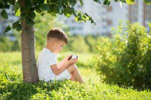 vrolijk jongen zittend Aan de gras looks tekenfilms in de telefoon in de zomer Bij zonsondergang. schattig baby hebben pret in natuur foto