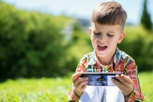 vrolijk kind zittend Aan de gras looks tekenfilms in de telefoon in de zomer Bij zonsondergang. schattig jongen hebben pret in natuur foto