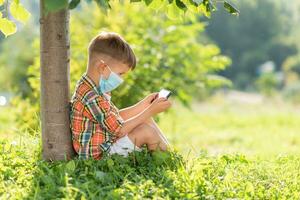 een kind in een medisch masker zit Aan de gras en looks in de telefoon tekenfilms in de zomer Bij zonsondergang. kind met een mobiel telefoon in zijn handen. het voorkomen tegen coronavirus covid-19 gedurende een pandemisch foto