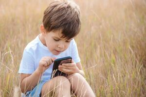 een knap jongen zittend Aan de gras Toneelstukken in de telefoon in de zomer spel Bij zonsondergang. de kind heeft pret in natuur foto