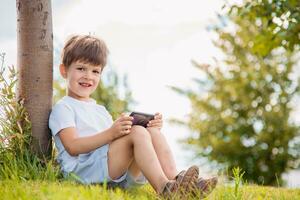 vrolijk kind zittend Aan de gras looks tekenfilms in de telefoon in de zomer Bij zonsondergang. schattig jongen hebben pret in natuur foto