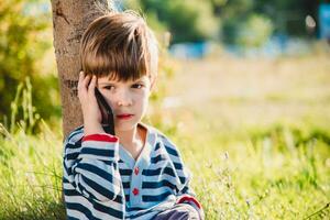 een mooi kind zittend Aan de gras spreekt door telefoon in de zomer Bij zonsondergang. jongen communiceert Aan mobiel foto