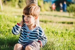 schattig jongen zittend Aan de gras spreekt door telefoon in de zomer Bij zonsondergang. de kind communiceert Aan een mobiel foto