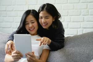 gelukkig Aziatisch lesbienne paar aan het doen online boodschappen doen Aan tablet zittend Aan sofa Bij huis. foto