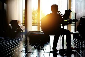 silhouet van een Mens zittend Aan een stoel spelen een elektrisch gitaar in hal ziekenhuis en achtergrond van de zonsondergang. silhouet romance concept, muziek- behandeling foto