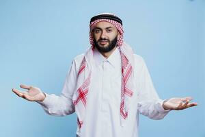 verward moslim Mens vervelend traditioneel kleren schouderophalend schouders en op zoek Bij camera met onzeker uitdrukking. Arabisch persoon gekleed in thobe verspreiden handen studio portret foto