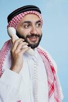 glimlachen Mens vervelend traditioneel moslim rood en wit geruit hoofdtooi chatten Aan vaste telefoon telefoon. vrolijk persoon in geruit Arabisch hoofdtooi met touw band hebben pret gesprek Aan telefoon foto