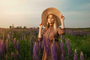 een mooi vrouw in een rietje hoed wandelingen in een veld- met Purper bloemen. een wandelen in natuur in de lupine veld- foto