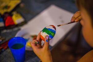 een kind siert een Pasen ei in de kleuren van de regenboog. een kind houdt een ei en verven het met een borstel. voorbereidingen treffen voor de viering van Pasen. foto