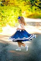 charmant klein meisje in een blauw lang jurk is dansen en wervelingen in een zonnig park. kind spinnen in een blauw rok. de weinig meisje wandelingen in de binnenplaats Aan een achtergrond van groen struiken lit door de zon. foto