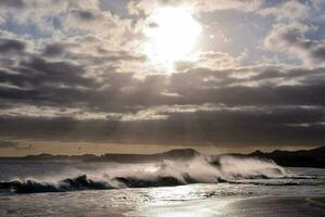 de zon schijnt door de wolken over- golven crashen Aan een strand foto