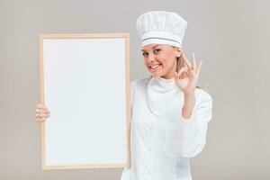 portret van mooi vrouw chef tonen OK teken terwijl Holding whiteboard Aan grijs achtergrond. foto