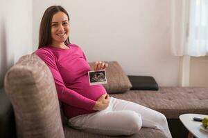 zwanger vrouw zittend Aan de sofa en Holding echografie afbeelding. foto