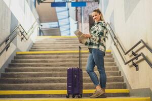 gelukkig vrouw met koffer op zoek Bij kaart terwijl staand Aan trap van de trein station foto