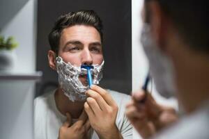 Mens scheren baard terwijl op zoek zichzelf in de spiegel foto