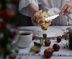 handen houden een croissant en gebruik een mes naar verspreiding aardbei jam Aan het en Aan de tafel is een kop van koffie en klein rood appels. selectief focus foto