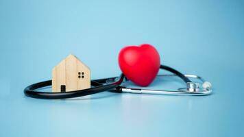 concept van Gezondheid verzekering en medisch welzijn, klein houten huis en rood hart met stethoscoop Aan blauw achtergrond, Gezondheid verzekering en toegang naar gezondheidszorg. foto