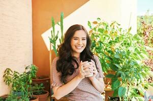 romantisch portret van mooi Aziatisch jong vrouw, ontspannende Aan de balkon tussen veel groen planten, Holding kop van thee of koffie foto