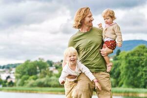 buitenshuis portret van gelukkig jong vader Holding twee kinderen in armen, kleuter meisje en kleuter jongen hebben pret met vader Aan een mooi hoor zonnig dag foto