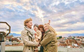 buitenshuis portret van gelukkig familie van vier, jong paar met twee weinig kinderen, verkoudheid het weer, oud Europese stad Aan achtergrond foto