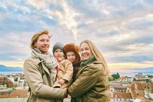 buitenshuis portret van gelukkig familie van vier, jong paar met twee weinig kinderen, verkoudheid het weer, oud Europese stad Aan achtergrond foto