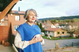 portret van mooi 50 -55 jaar oud vrouw poseren Aan balkon, Holding kop van thee of koffie foto