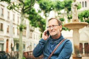 buitenshuis portret van midden- leeftijd Mens pratend Aan de telefoon, vervelend bril en blauw trui, Holding smartphone De volgende naar oor foto