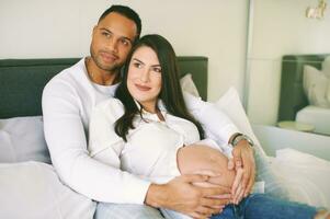 binnen- portret van gelukkig jong familie, zwanger vrouw met haar liefhebbend man ontspannende Aan bed foto