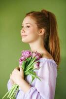 schoonheid portret van mooi jong 15 - 16 jaar oud roodharige tiener meisje vervelend Purper jurk poseren Aan groen achtergrond, Holding tulp bloemen foto