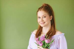 schoonheid portret van mooi jong 15 - 16 jaar oud roodharige tiener meisje vervelend Purper jurk poseren Aan groen achtergrond, Holding tulp bloemen foto