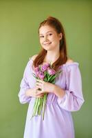 schoonheid portret van mooi jong 15 - 16 jaar oud roodharige tiener meisje vervelend Purper jurk poseren Aan groen achtergrond, Holding tulp bloemen foto