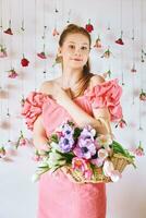 studio portret van mooi jong tiener- 15 - 16 jaar oud roodharig meisje vervelend roze koraal jurk, poseren Aan wit achtergrond met hangende rozen, Holding mand met veel voorjaar bloemen foto