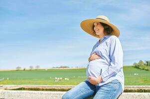 buitenshuis portret van gelukkig jong zwanger vrouw genieten van mooi hoor dag in platteland, zittend Aan gieter fontein, groen weiland met koeien Aan achtergrond foto