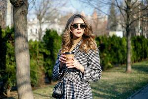 vrouw in loopgraaf jas wandelen en drinken koffie van papier kop foto