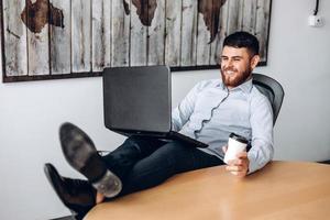 aardige vent met een baard die aan een tafel zit, zijn voeten op tafel zet, koffie drinkt en op een computer werkt foto