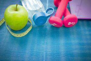 twee rood halters, een appel, een plakband meeteenheid, een fles van water en gewicht balans Aan een yoga mat. concepten over fitheid, sport en Gezondheid foto