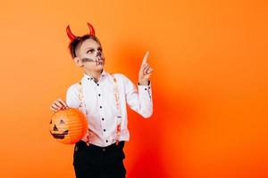 jongen in duivelsmaskerademake-up die een pompoen houdt en omhoog wijst. halloween-concept foto