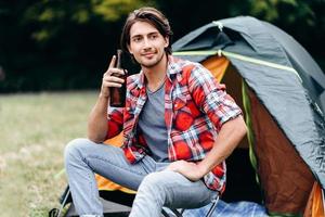 een man die een fles bier vasthoudt en glimlachend naar buiten kijkt. tent op de achtergrond foto