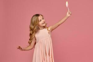 weinig meisje met een blond gekruld haar, in een roze jurk is poseren met een snoep foto