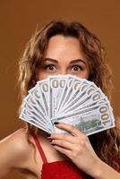 beeld van Lucky gelukkig vrouw met bruin lang haar- met ventilator van 100 dollar rekeningen, veel van contant geld geld, over- bruin achtergrond foto